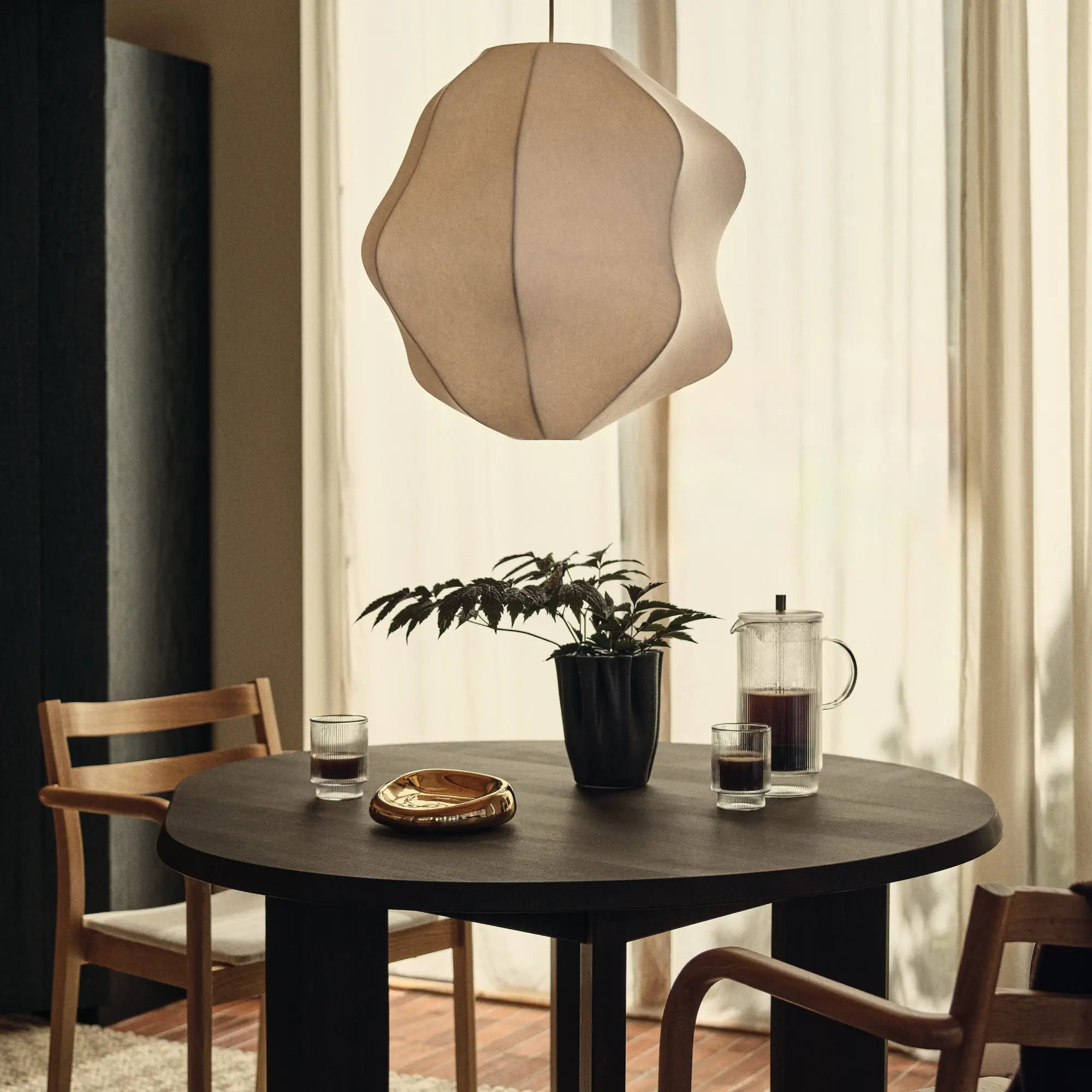 Matrummet, vågig taklampa i tyg ovanför ett runt mörkt matbord med glas, en kaffepress och en växt