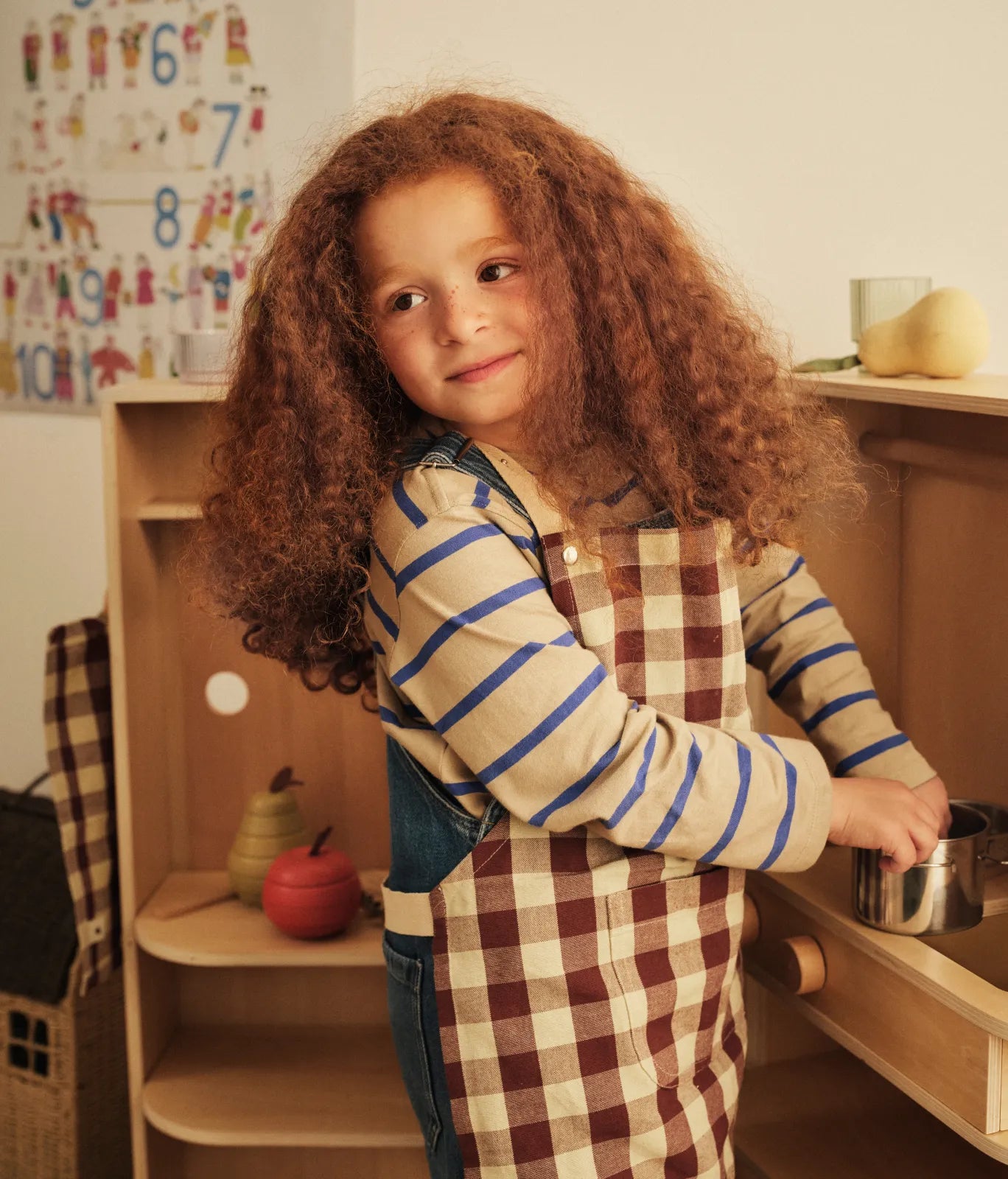 Kids Autumn Winter 2026, barn i rutigt förkläde håller en kastrull vid ett lekkök, leksaksfrukt på hyllan