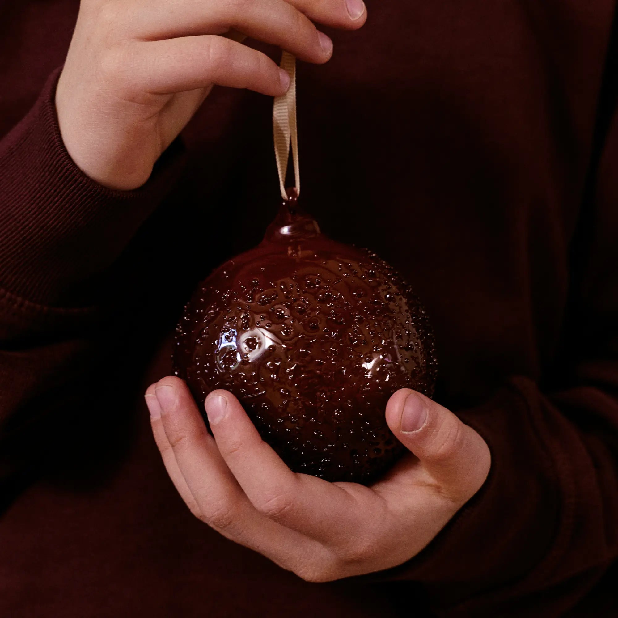 Julkollektionen, händer som håller en julkula i bubbligt glas i mörkrött med en ögla av band