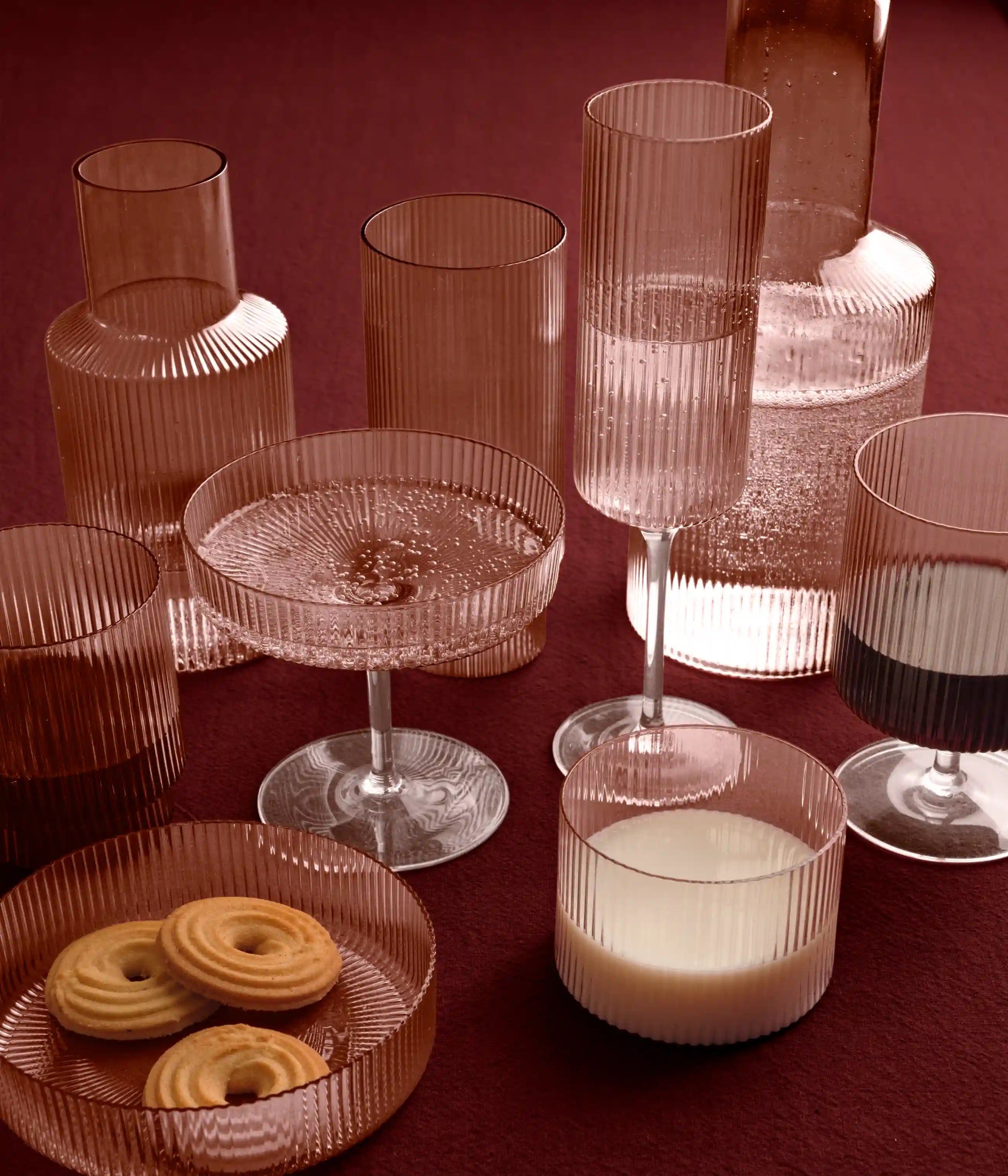 Räfflat glas i rosa på ett mörkrött bord, karaff, glas och ett fat med kex