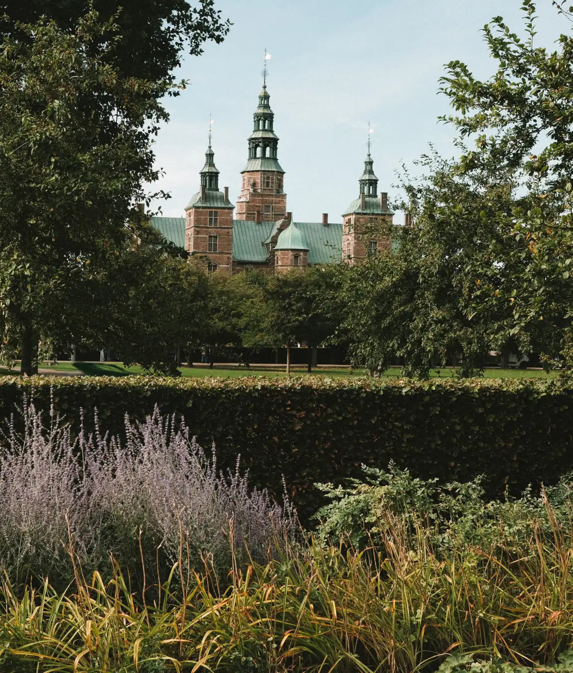 Slott med gröna spiror sett över en park, häck och blommande gräs i förgrunden