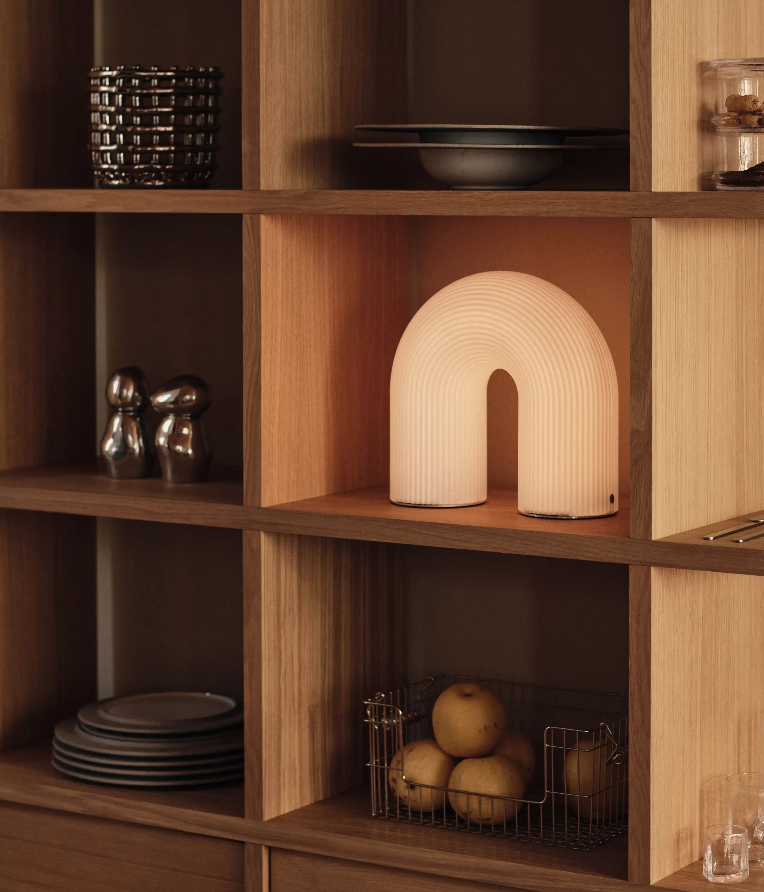 Tänd bågformad lampa på en hyllmodul med tallrikar, skålar och en trådkorg med frukt