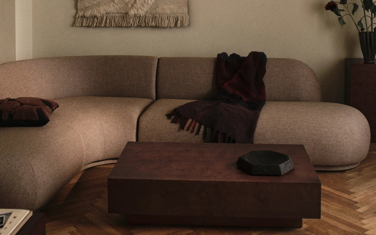 Böjd soffa i beige med ett mörkt pläd och lågt soffbord på fiskbensparkett