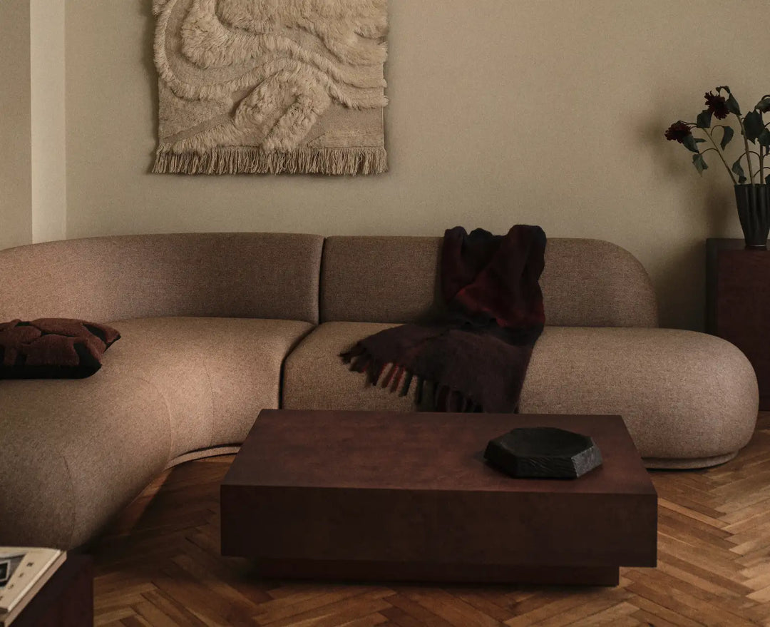 Böjd soffa i beige med ett mörkt pläd och lågt soffbord på fiskbensparkett