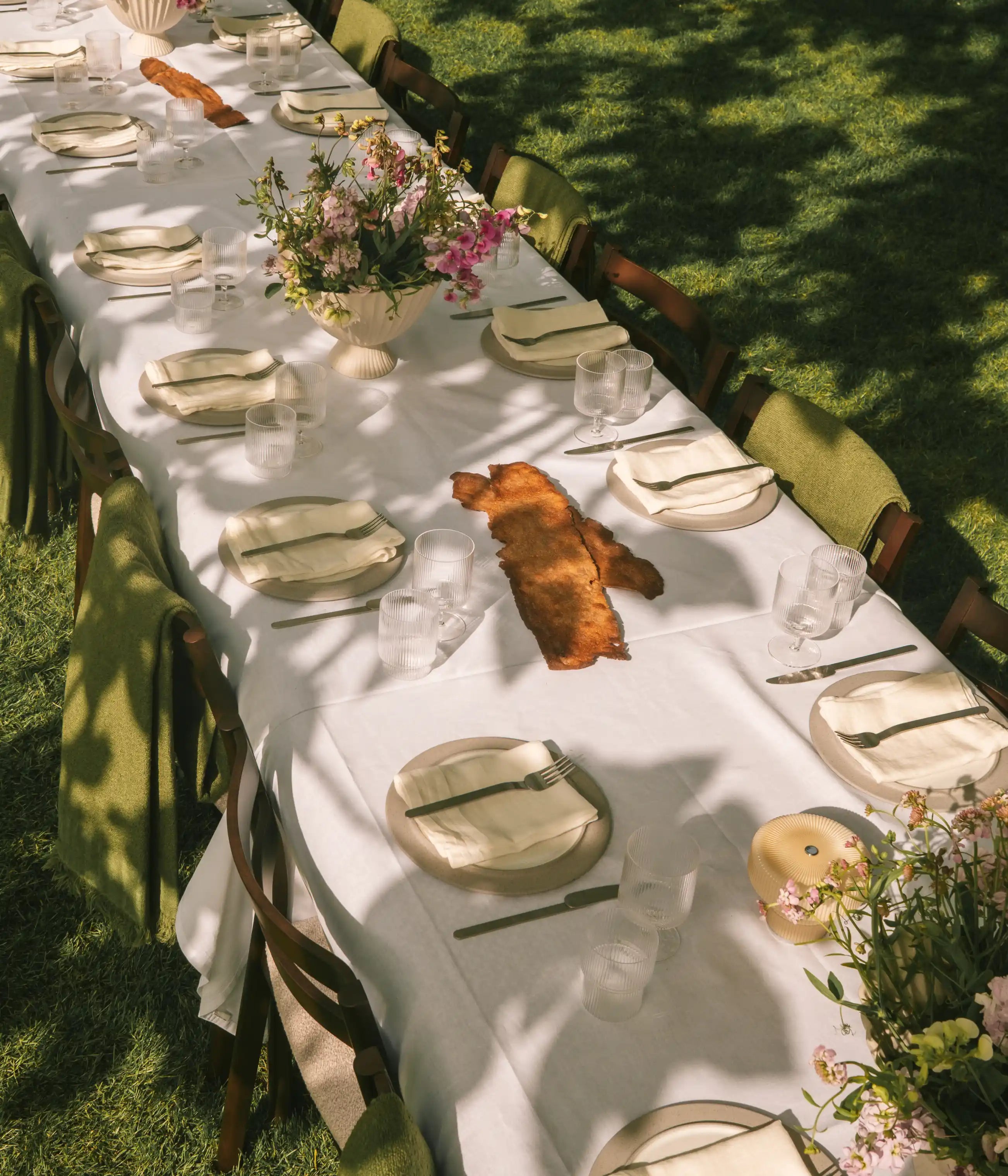 Utemöbler, långt bord dukat på en gräsmatta med vit duk, räfflade glas, blommor och gröna plädar på stolarna